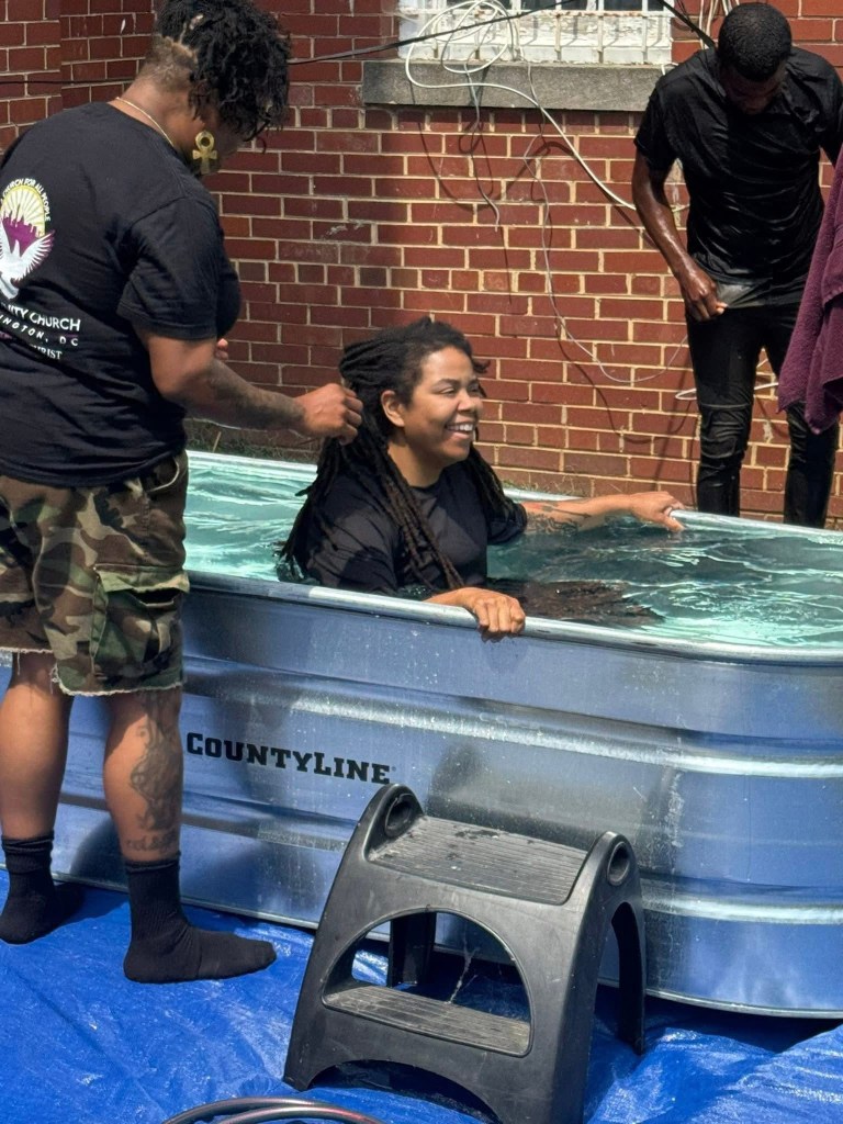 A woman with long hair is sitting in a water-filled metal tub, smiling as another person stands behind her, holding her hair. A third person is standing nearby, both dressed in black clothing. The scene is outdoors, with a brick building in the background.